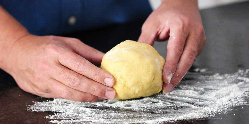 Two hands form pasta dough into a ball on a floured work surface. 