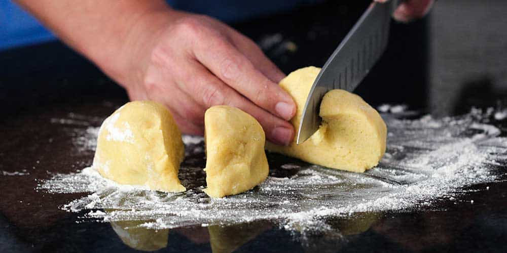 A knife cutting homemade pasta dough into 4 pieces on a floured work surface. 