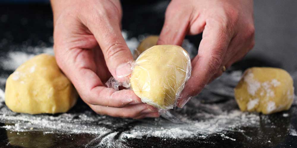 Two hands wrapping a piece of homemade pasta dough in plastic wrap. 