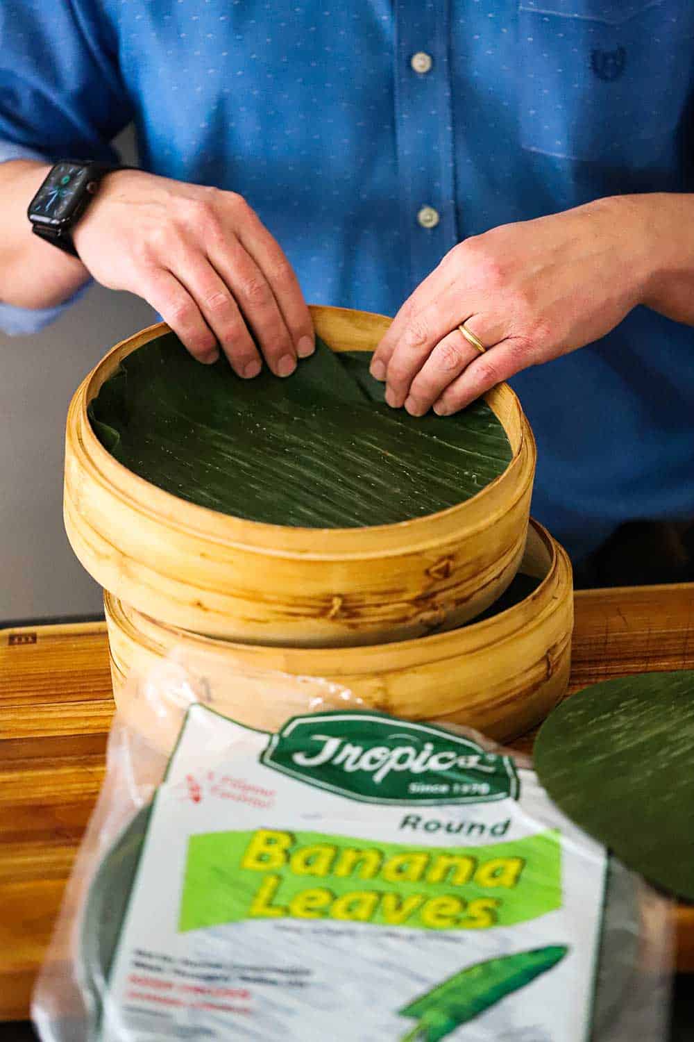 A person placing a circular piece of banana leaves into a double-stacked bamboo steamer with the package of banana leaves in the foreground. 