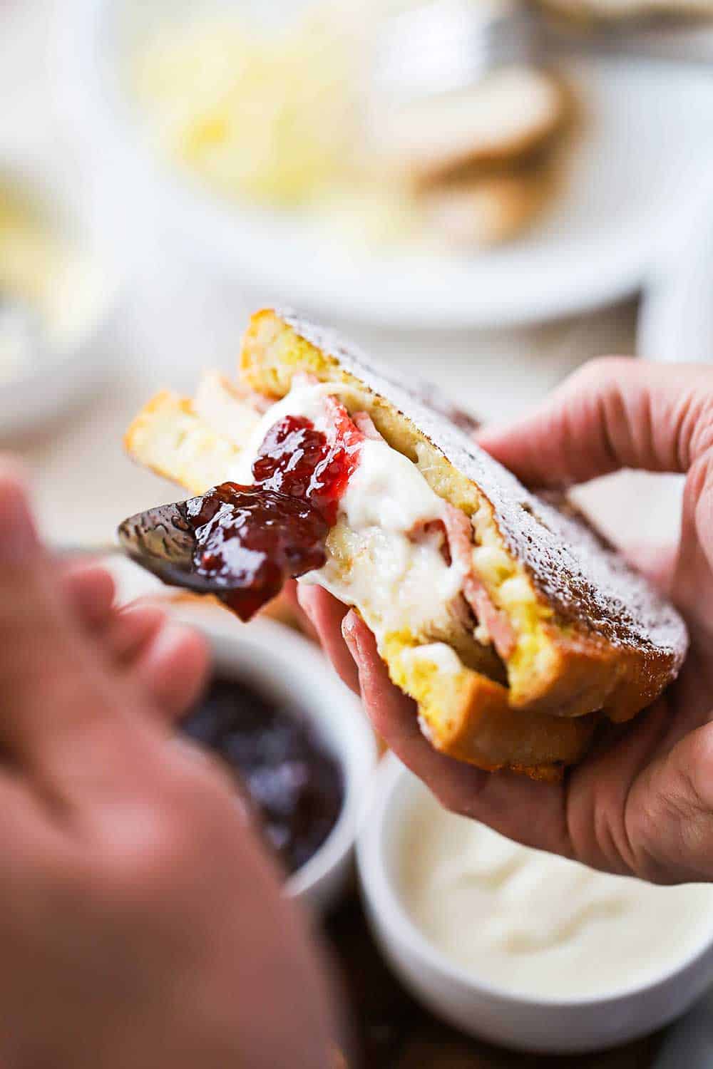 A person using a spoon to spread preserves along the cut side of a monte cristo sandwich. 