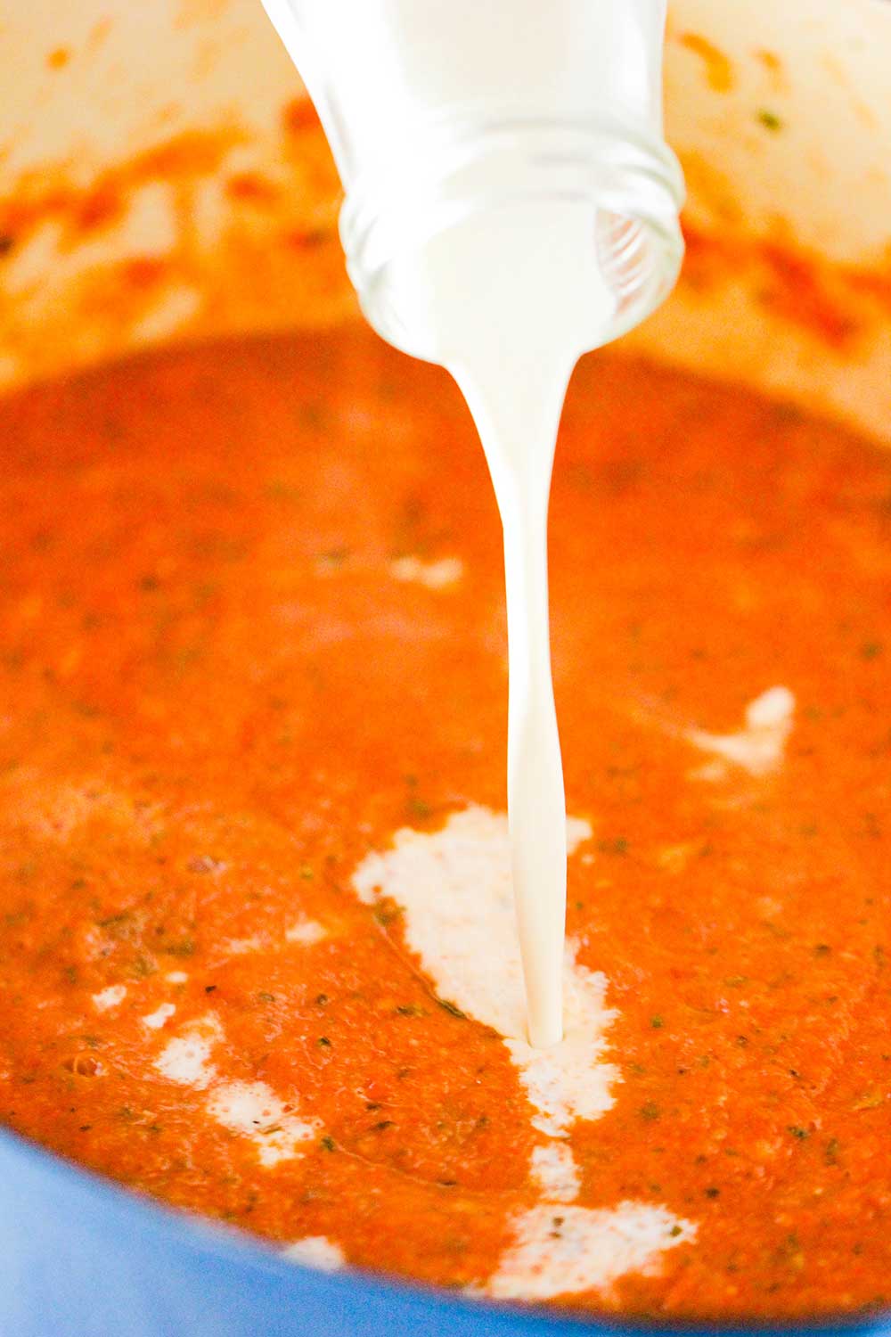 Heavy cream being poured from a milk jar into a pot of tomato soup. 