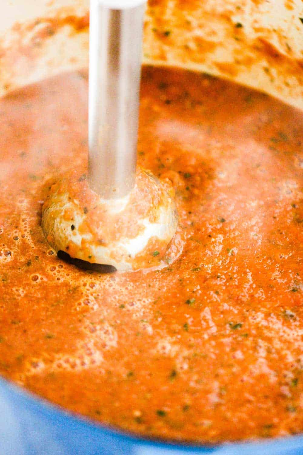 An immersion blender puréeing roasted tomato basil soup in a large pot. 