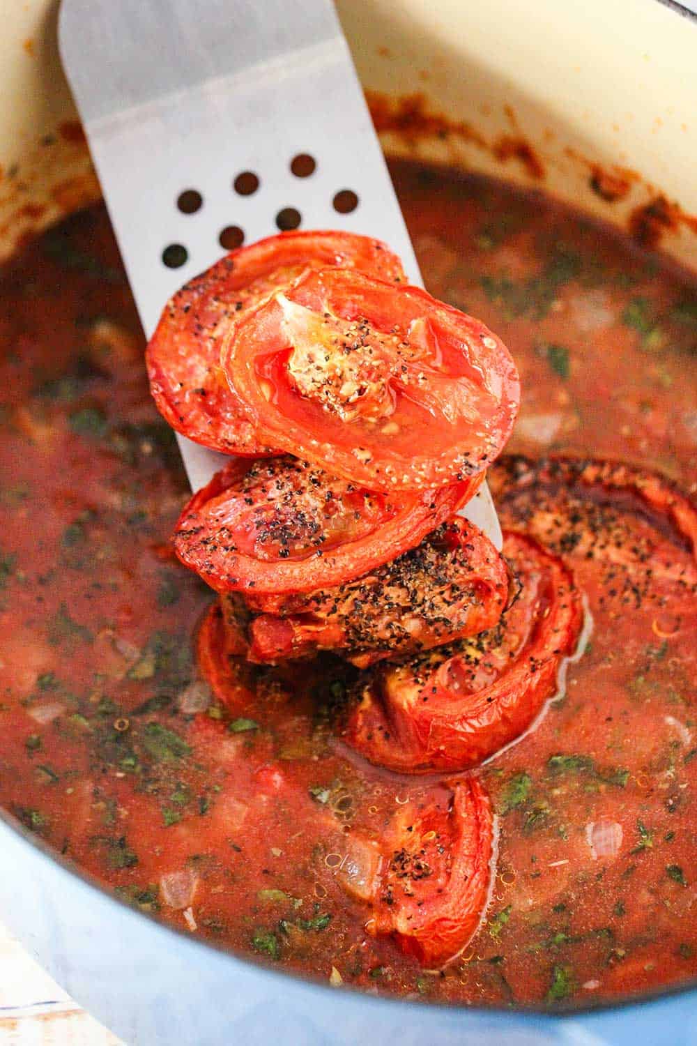 A metal spatula dropping roasted tomatoes into a pot of tomato basil soup. 