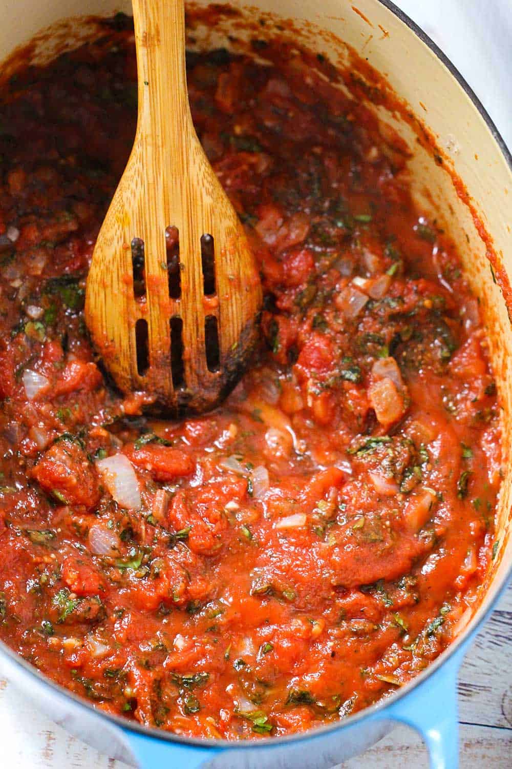 A large blue Dutch oven with tomatoes and herbs simmering in it with a wooden spoon in the pot. 