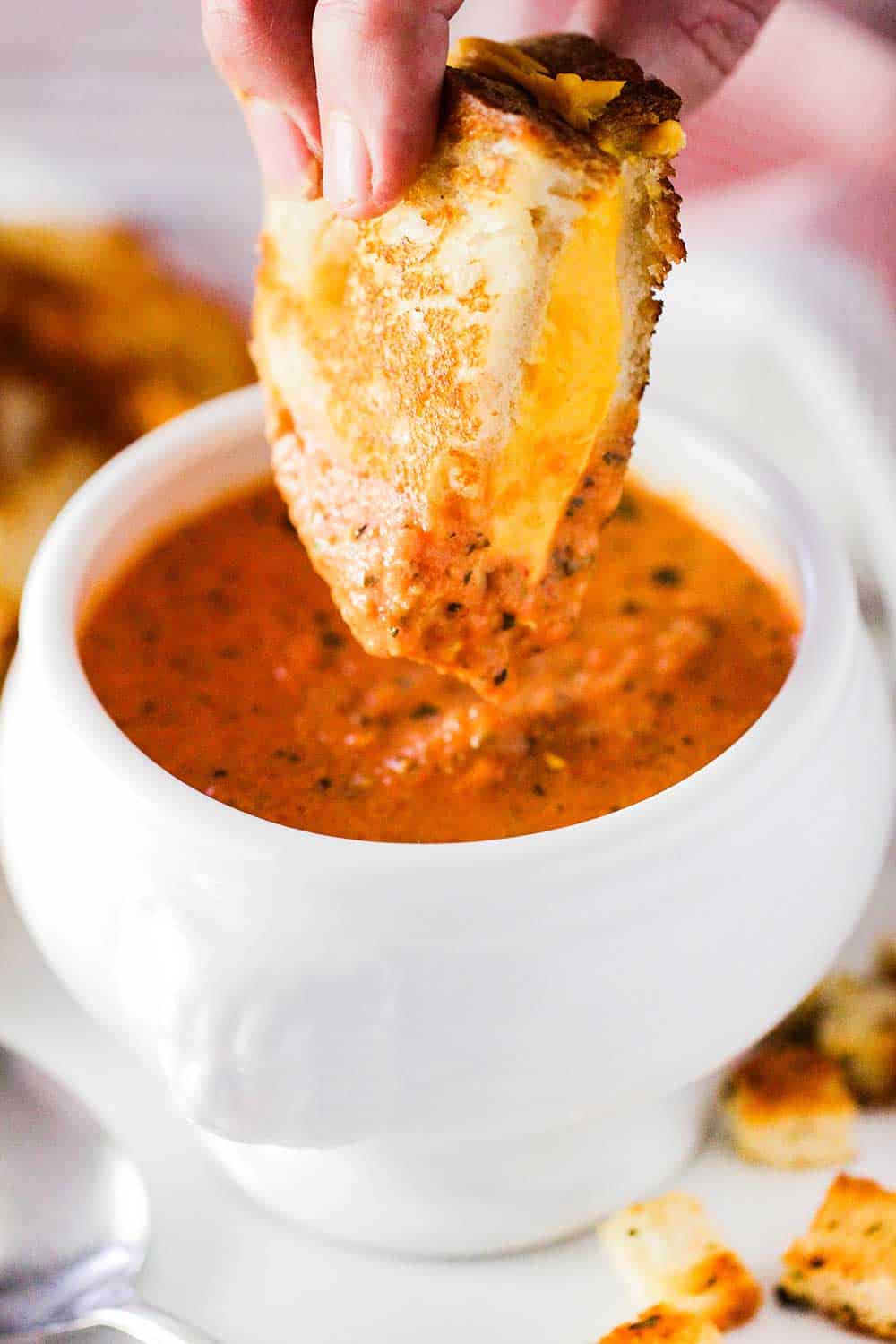 A hand dunking a grilled cheese sandwich into a bowl of tomato basil soup. 