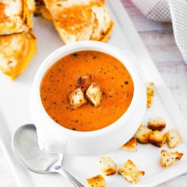 A white soup filled with roasted tomato basil soup next to sliced grilled cheese sandwiches.