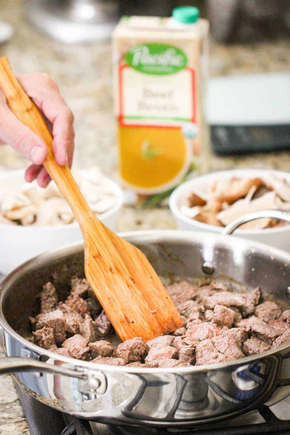 A wooden spoon stirring cooked beef pieces in a large silver skillet on a stove.