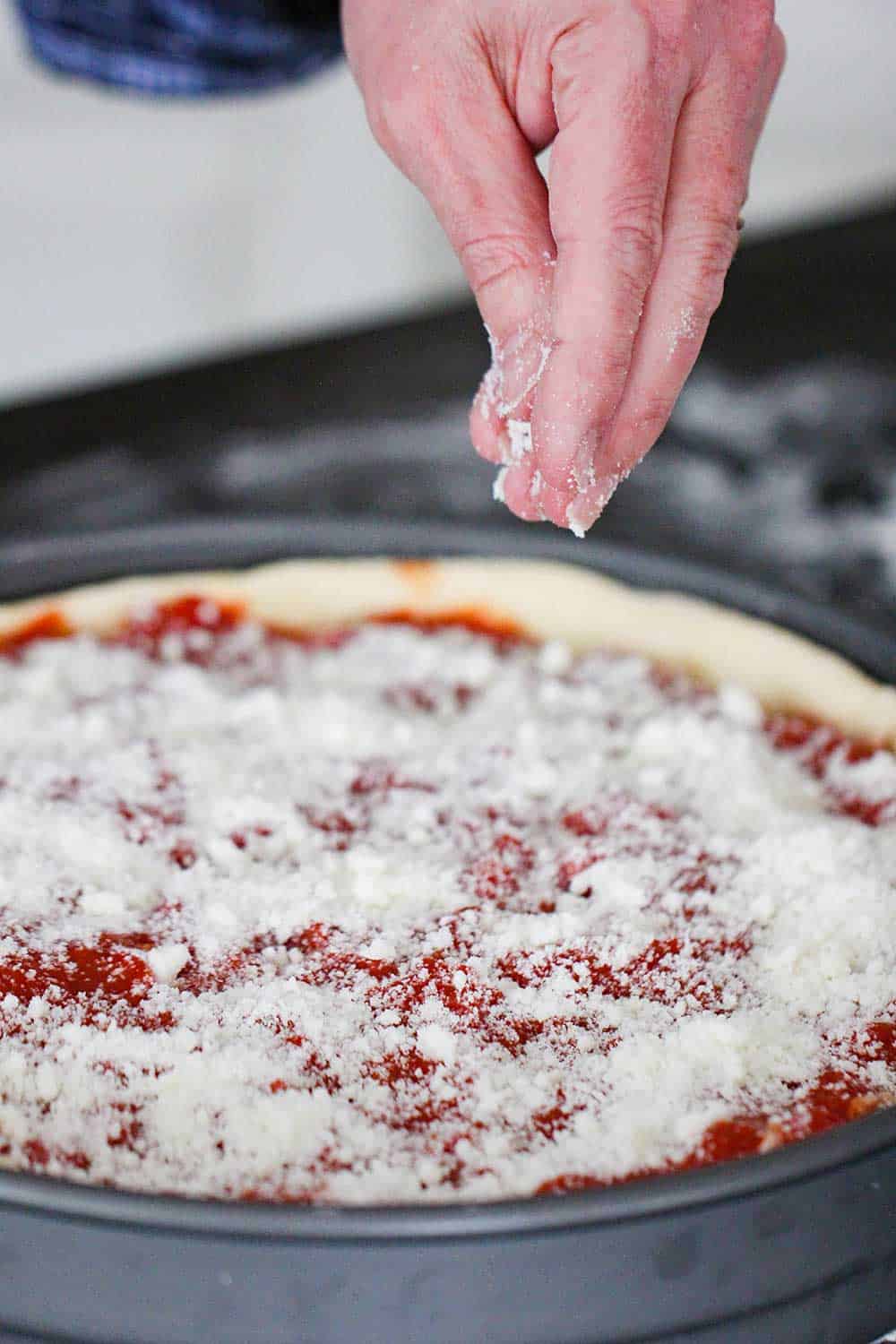 Fingers sprinkling Parmesan cheese over an unbaked Chicago-Style Deep Dish Pizza. 