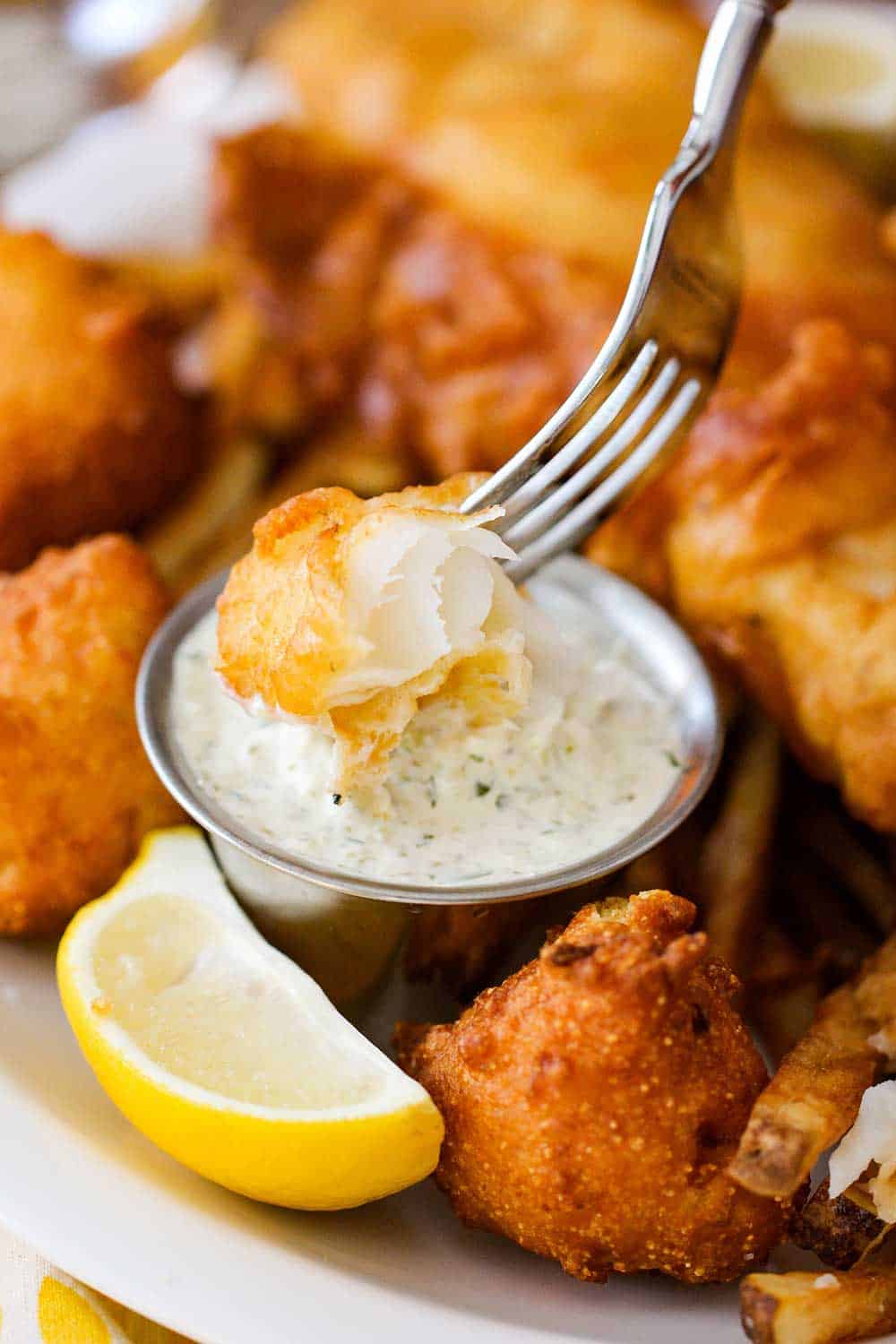 A piece of fried fish on a fork being dipped into a small bowl of homemade tartar sauce next to a lemon wedge. 