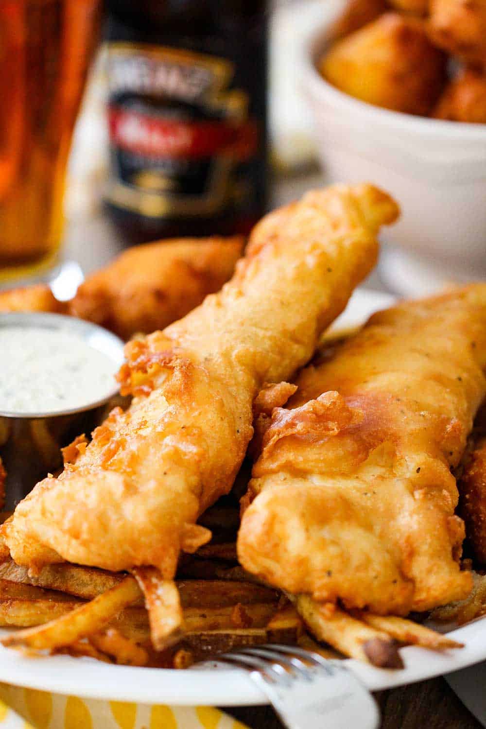 A plate of fish and chips with a small bowl of homemade tartar sauce and a bowl of hush puppies and a beer in the background. 