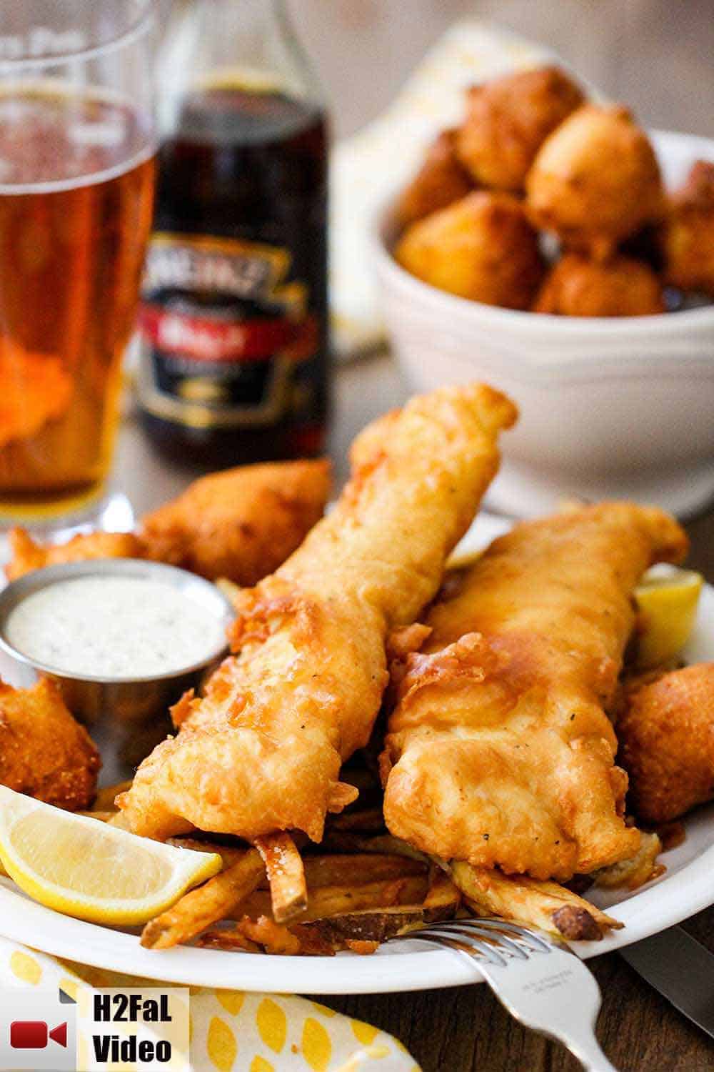 A plate of English fish and chips with a bowl of hush puppies and a glass of beer in the background. 