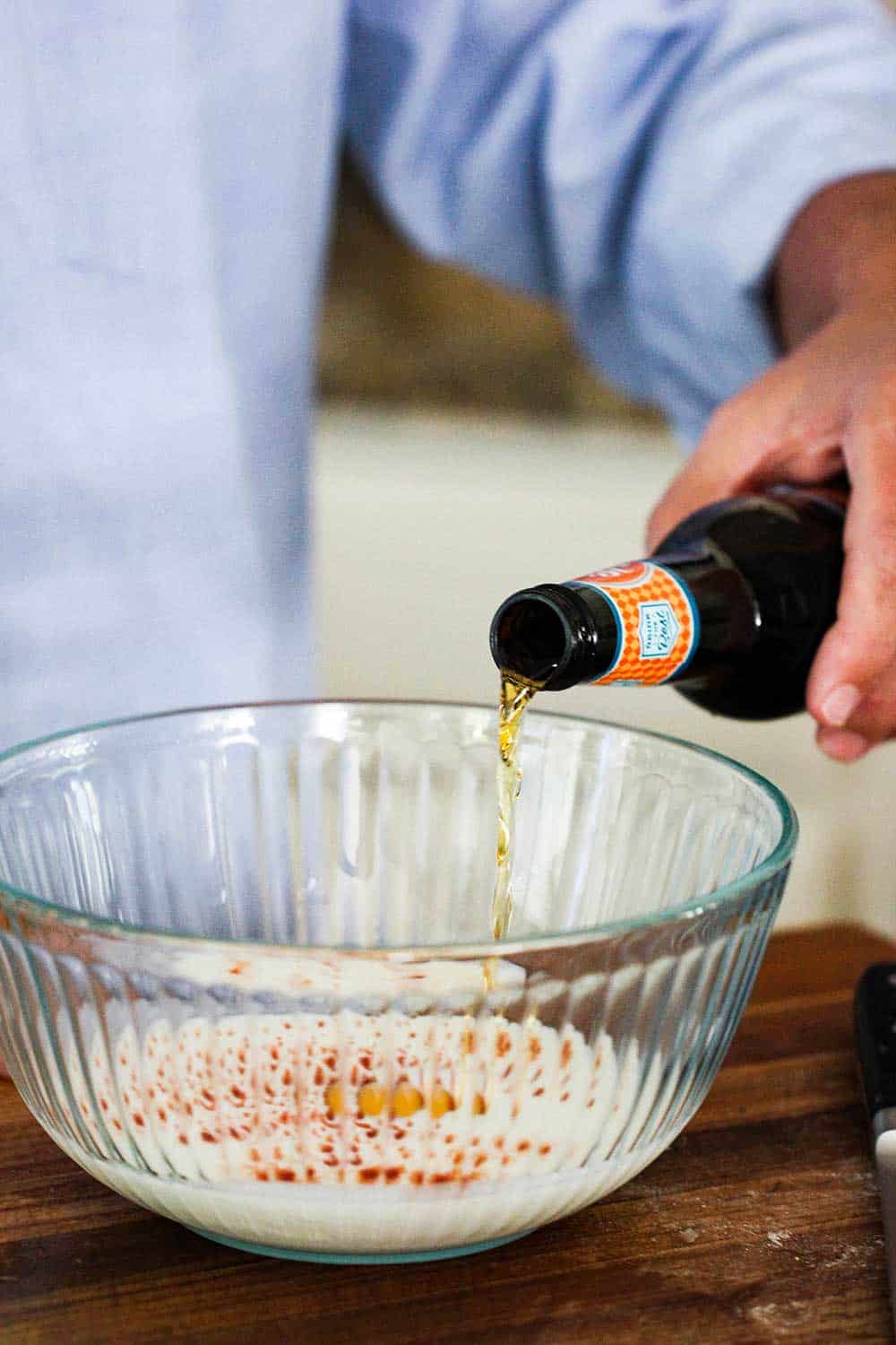 A person pouring a beer from a bottle into a bowl of hush puppies batter. 