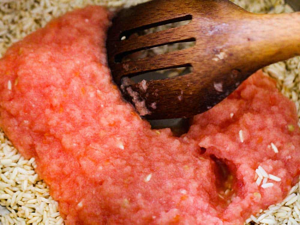 A wooden spoon in a pan with cooked rice and puréed tomatoes on top of it. 