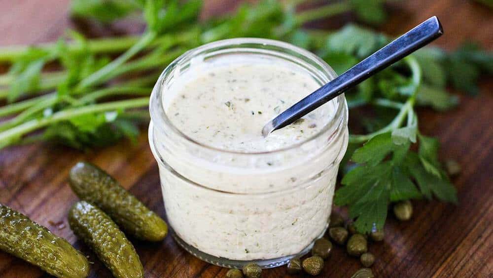 A small jar of homemade tartar sauce with pickles and fresh parsley next to it. 