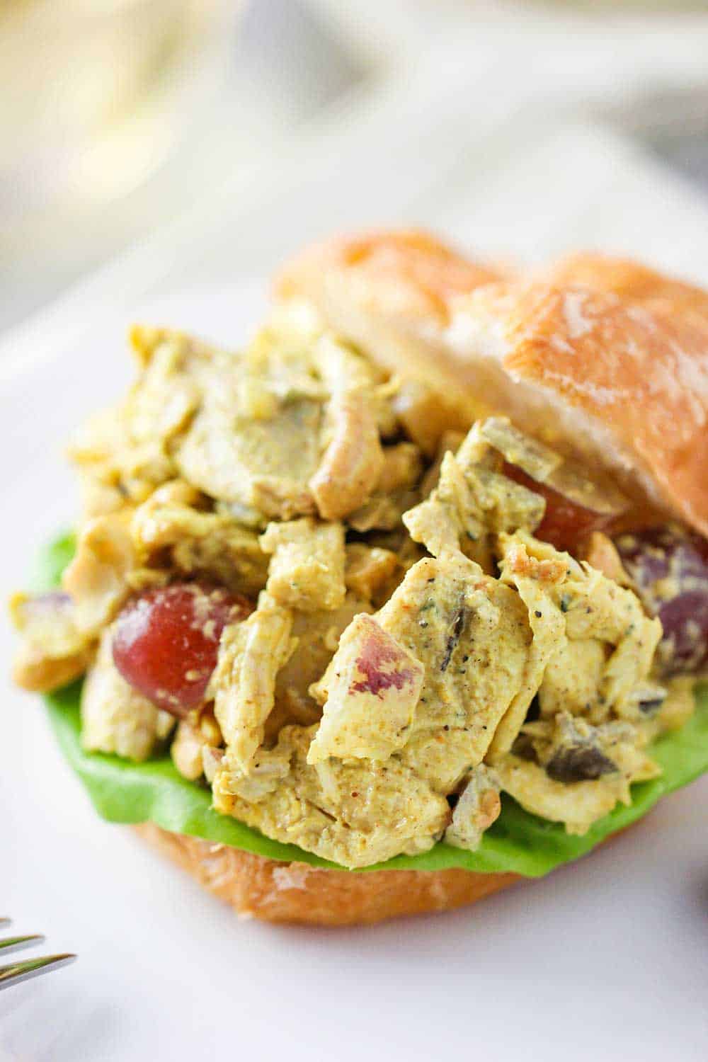 A close-up view of curry chicken salad between two croissant halves on a white plate. 