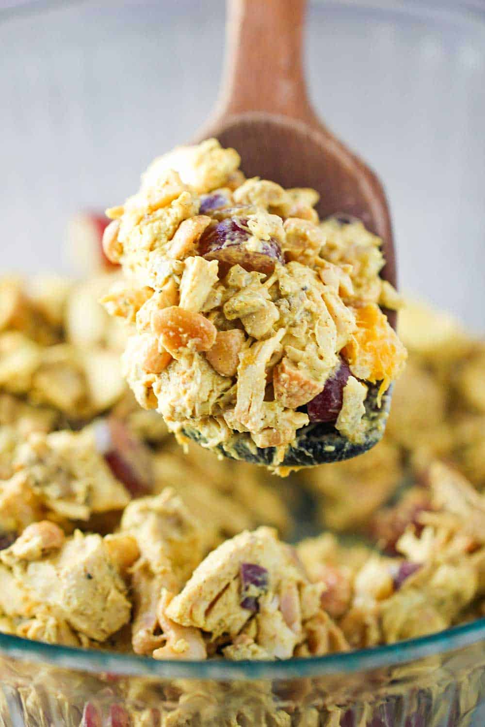 A large wooden spoon holding a heaping amount of curry chicken salad over a bowl filled with the mixture. 