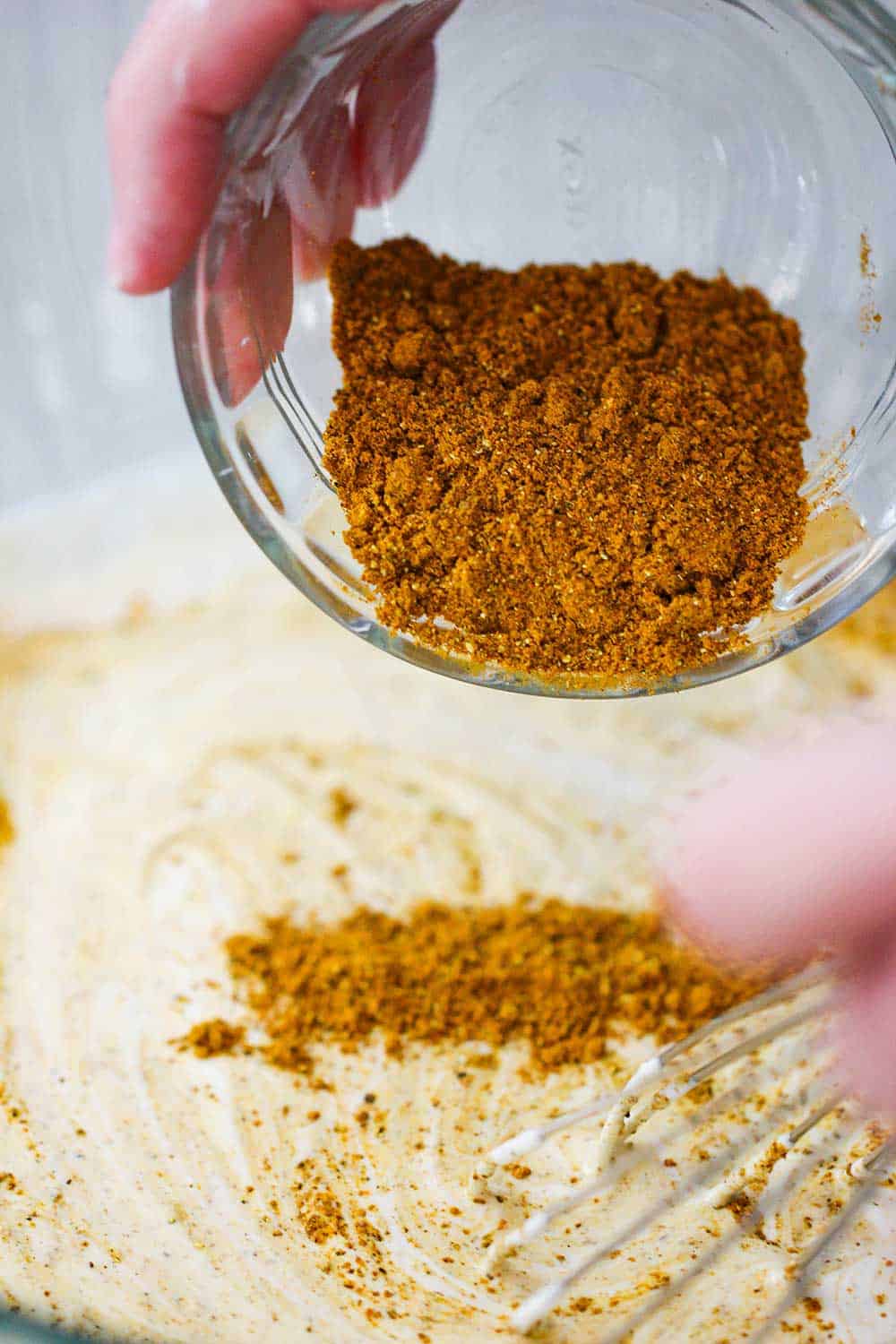 A hand sprinkling curry powder from a small glass bowl into another glass bowl of curry chicken salad dressing. 