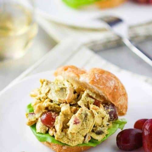 A croissant sandwich with curry chicken salad on a white plate next to a glass of white wine.