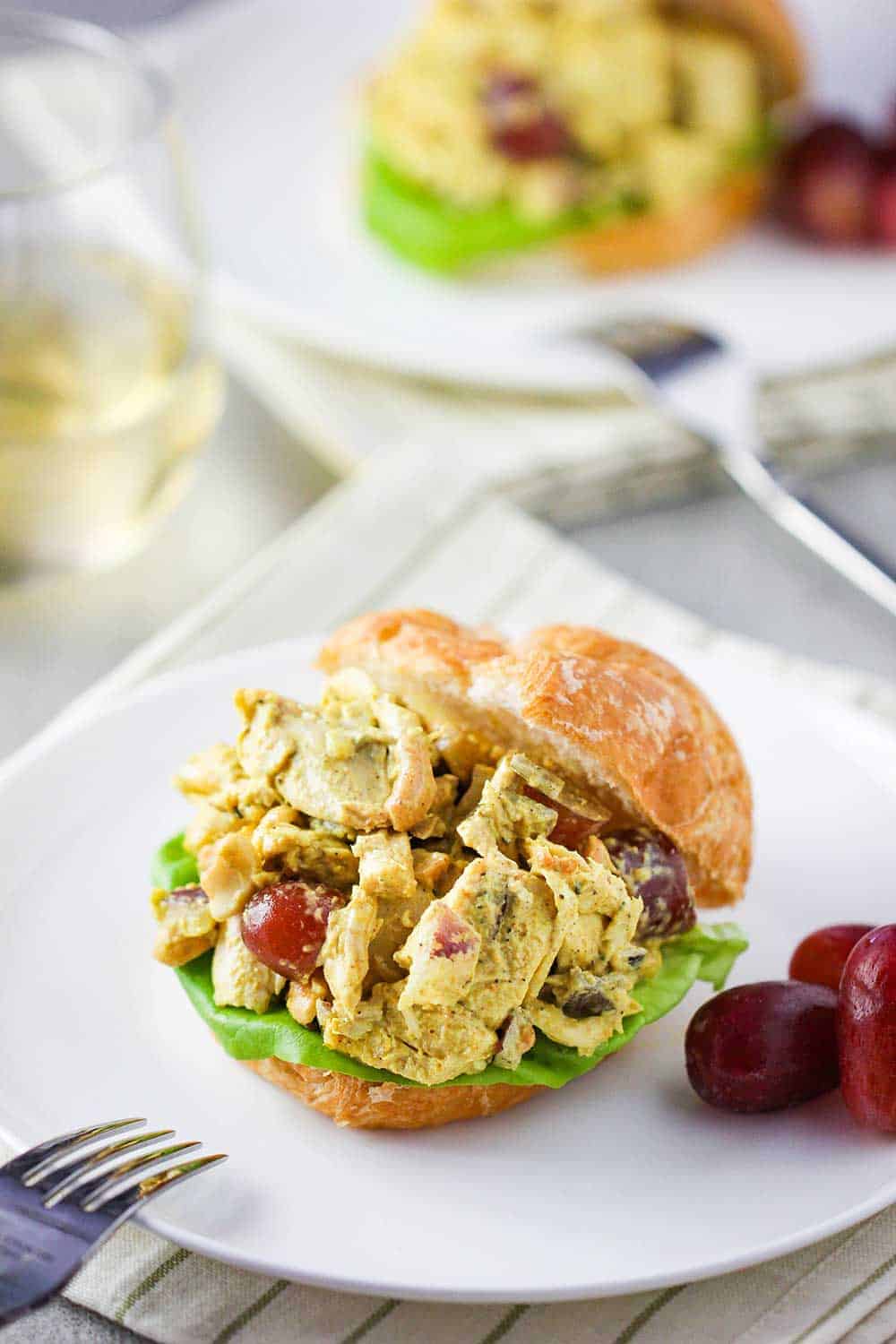 A croissant sandwich with curry chicken salad on a white plate next to a glass of white wine.