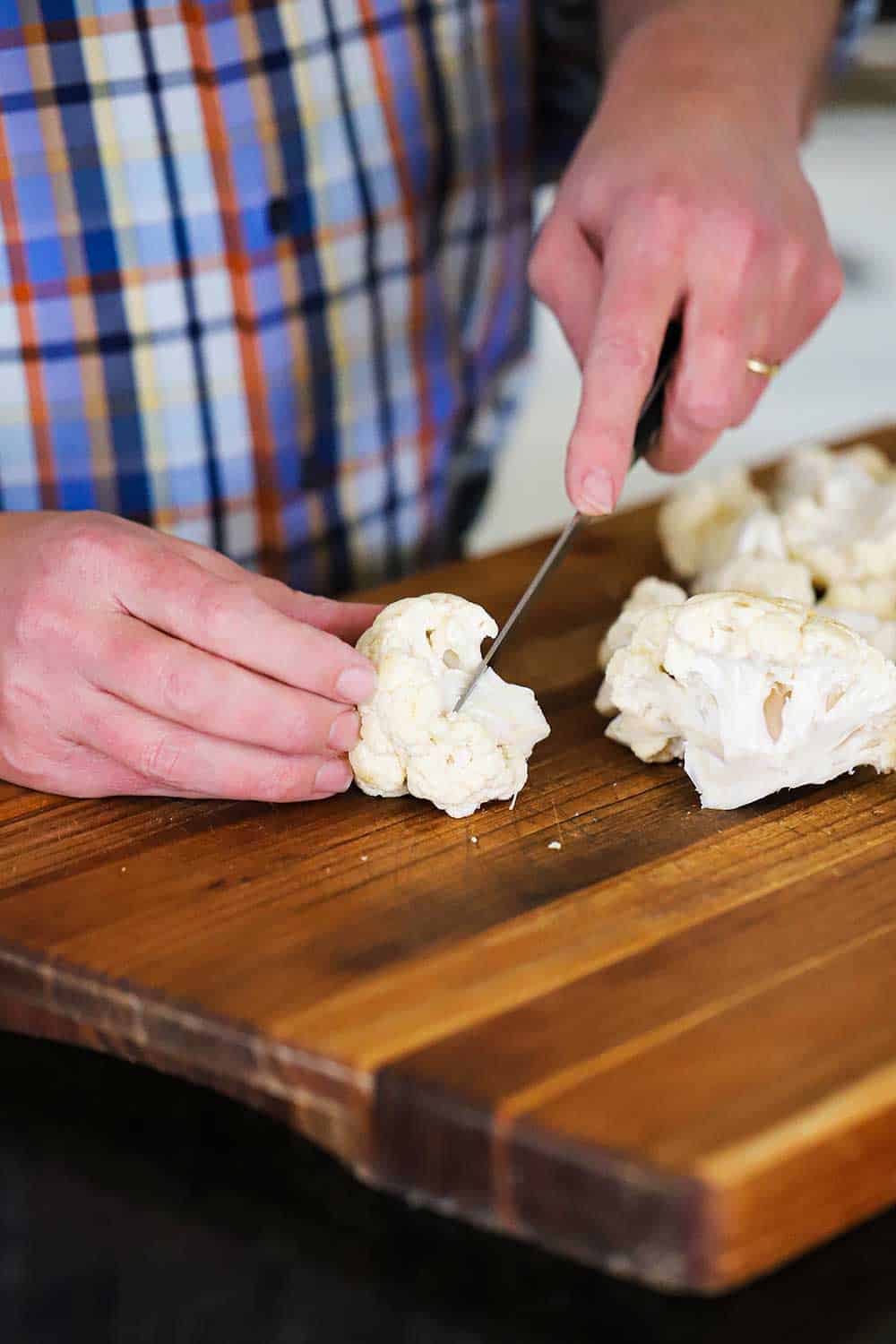 A person using a knife to cut a cauliflower heat into florets on a wooden cutting board. 