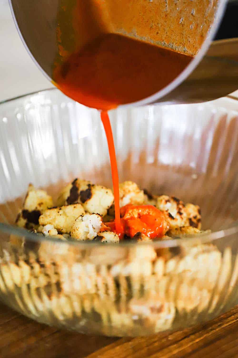 Buffalo sauce being poured from a silver saucepan into a glass bowl filled with roasted cauliflower pieces. 