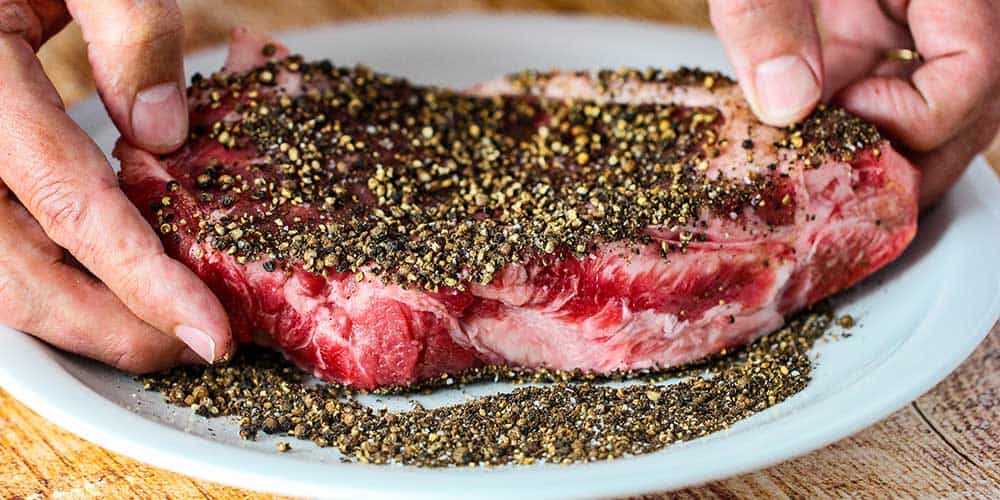 Spread the uncooked strip steaks in the cracked black peppercorns. 