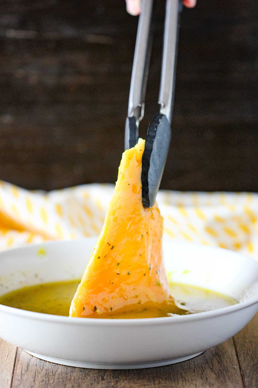 A pair of tongs lifting a thin uncooked chicken cutlet out of a bowl of honey mustard marinade. 