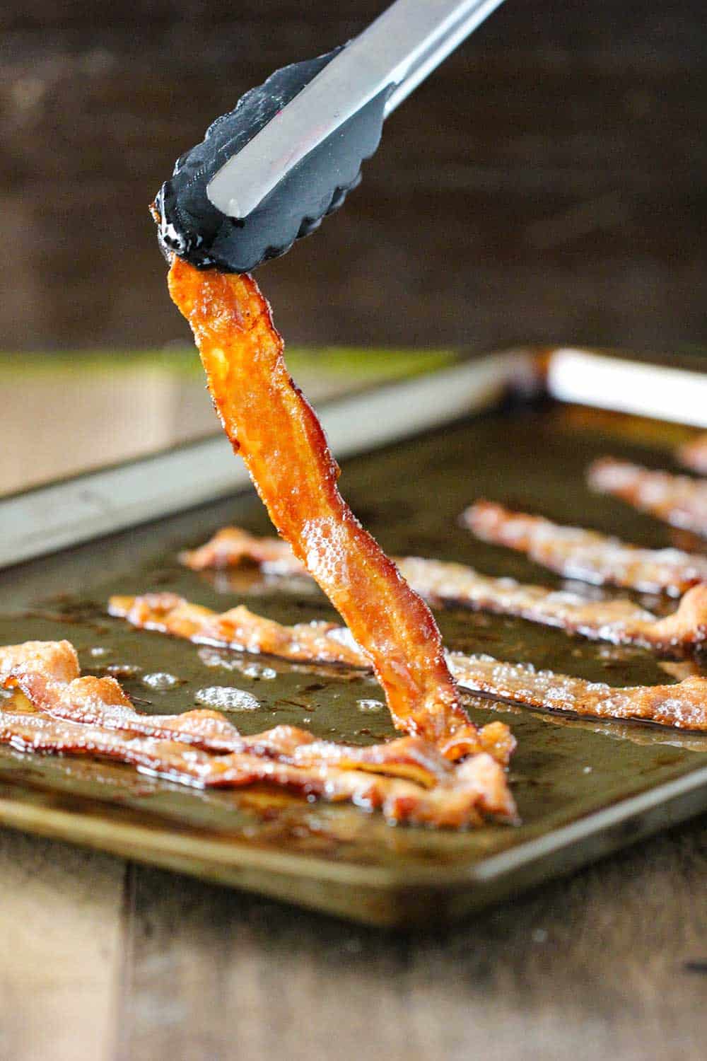 A pair of tongs lifting up a strip of cooked bacon from a baking sheet. 