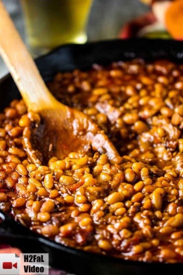 Stir a batch of cooked Southern Baked Beans with a wooden spoon in a large cast iron skillet.