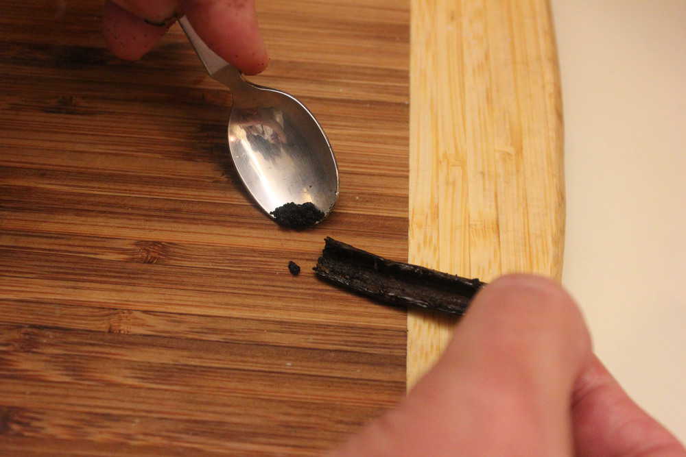 A hand using a spoon to scrape out the inside of a vanilla bean. 