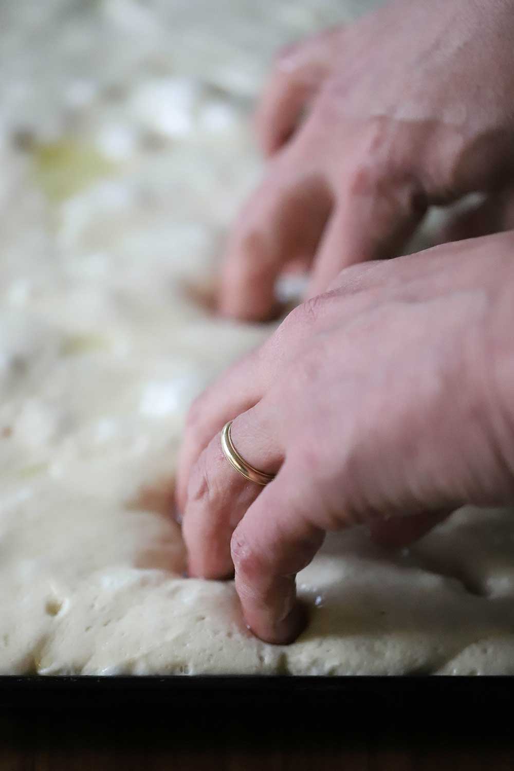 A pair of hands with fingers being pressed into bread dough to create deep dimples. 