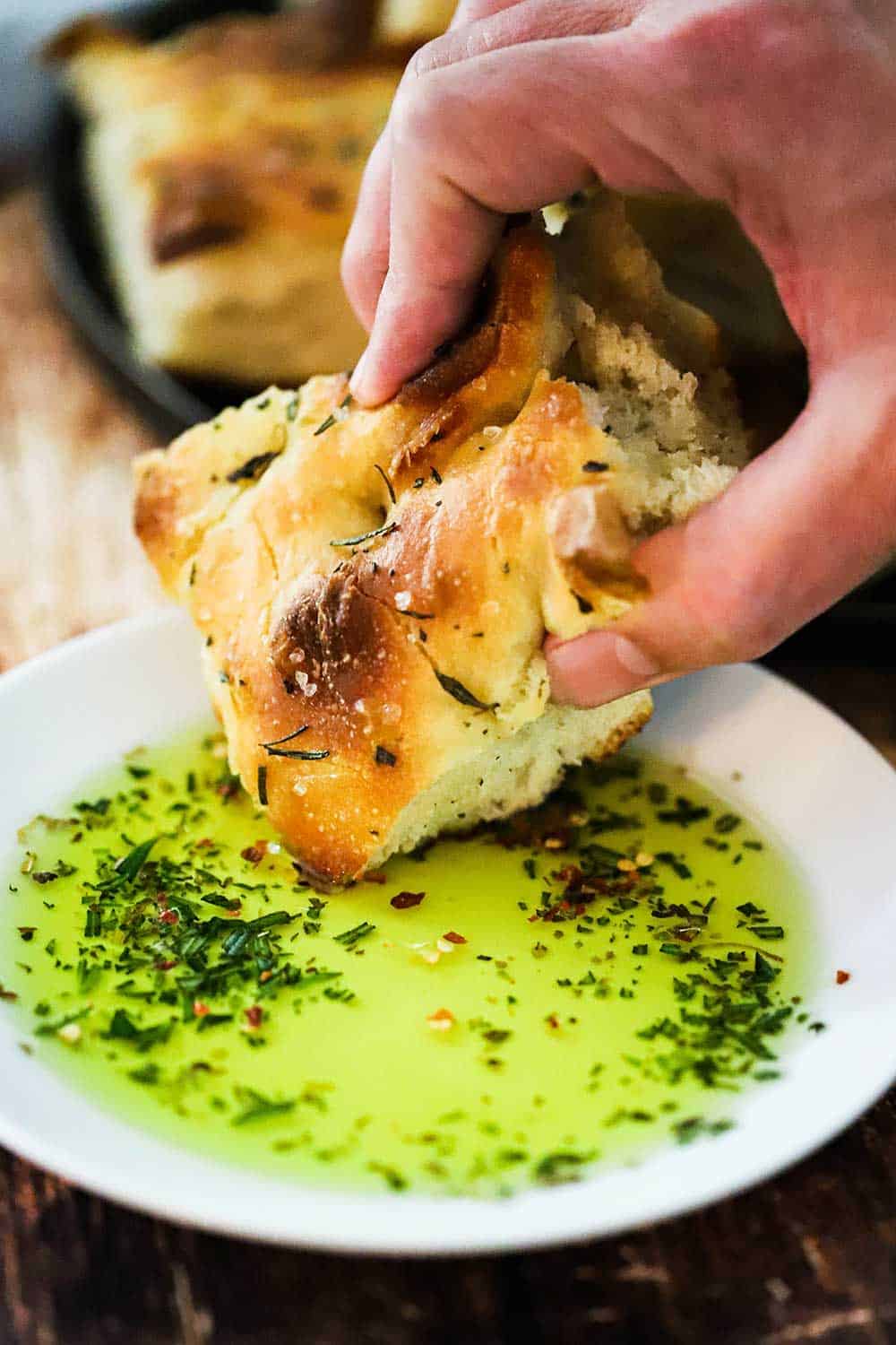 A hand dipping a square of focaccia bread into a plate that is filled with herbed olive oil. 