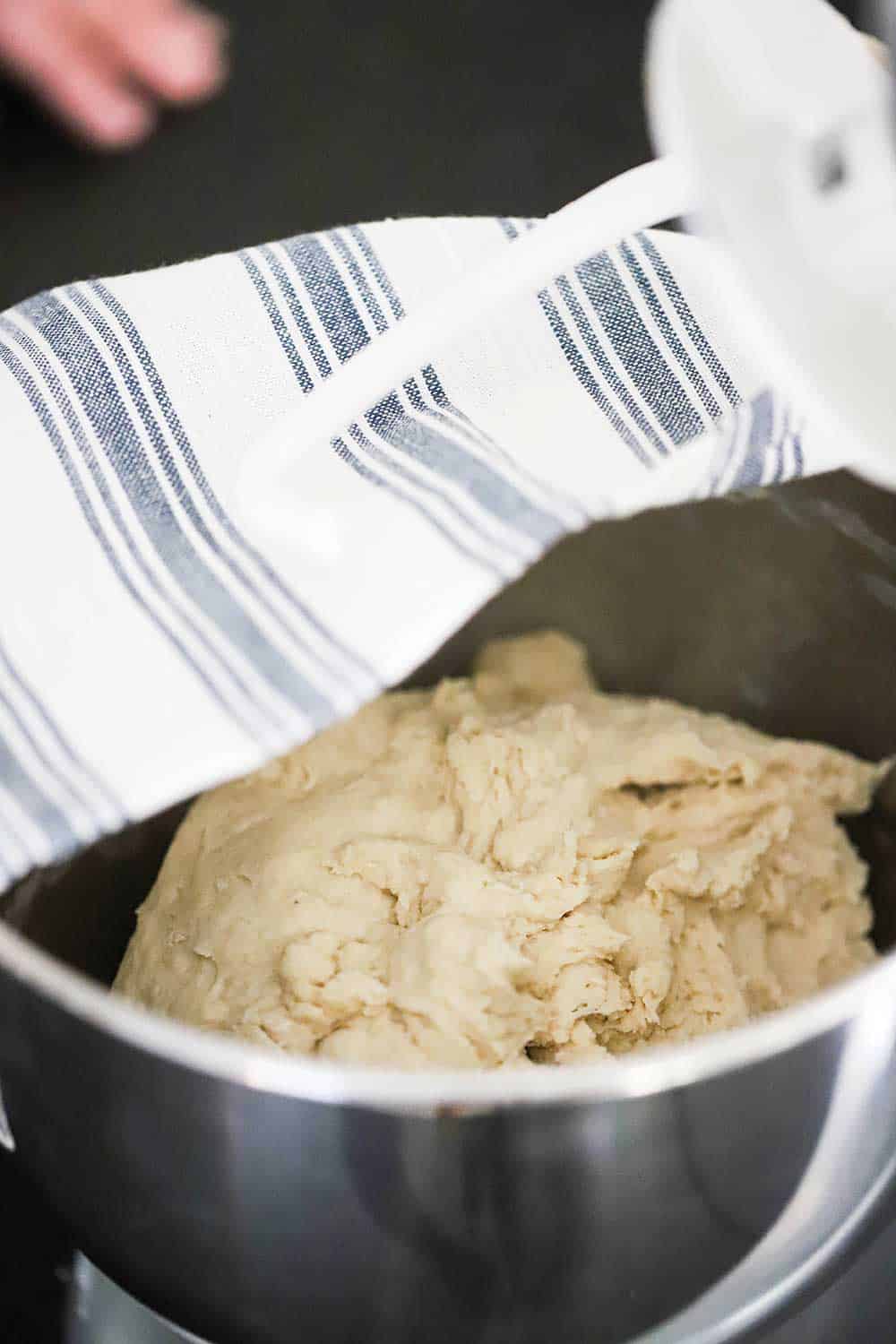 Bread dough in the bowl of a stand mixer with a striped kitchen towel being placed over the top of the bowl. 