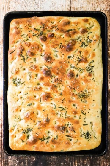 A large rectangular shaped loaf of focaccia bread with olive oil and fresh rosemary on top of it, all in a large rimmed baking pan.