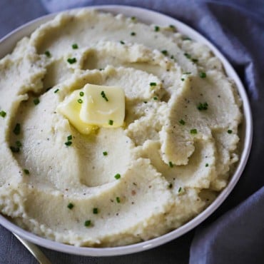 A white bowl filled with mashed cauliflower and roasted garlic with a pad of butter melting on top, all sitting next to a blue napkin and gold spoon.