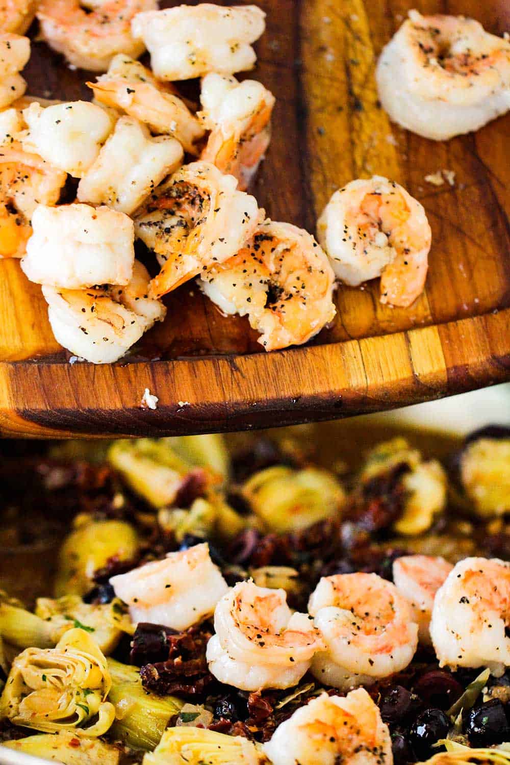 Seared shrimp falling from a wooden cutting board into a pan of cooked artichokes and olives. 