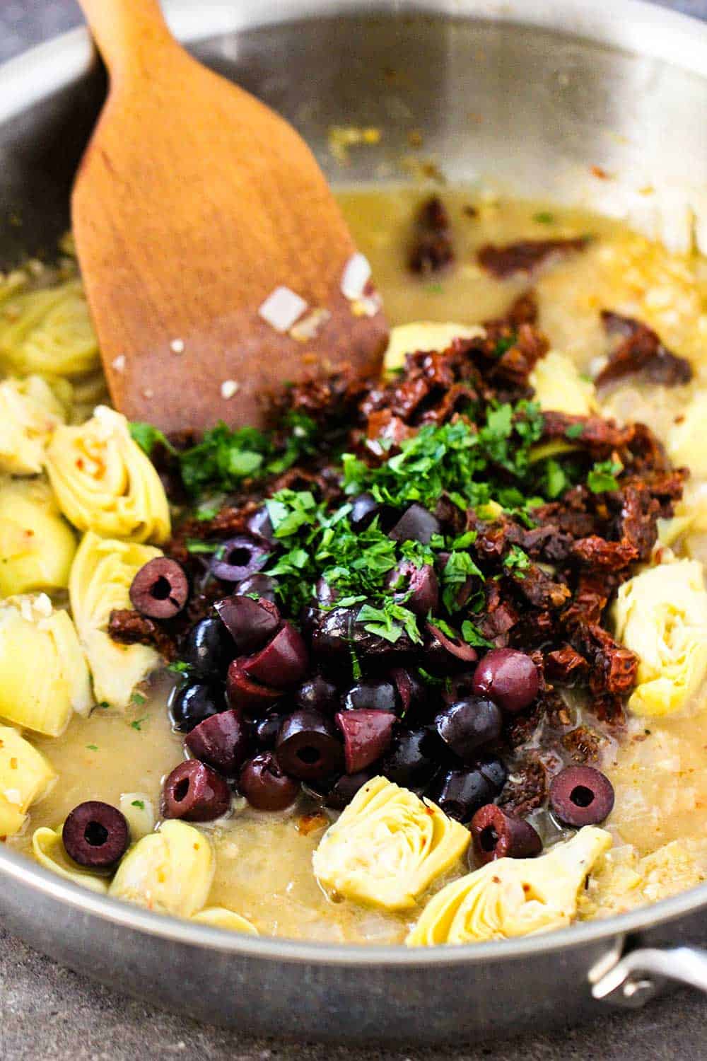 A large silver saucepan filled with olives and sun-dried tomatoes with a wooden spoon in it. 