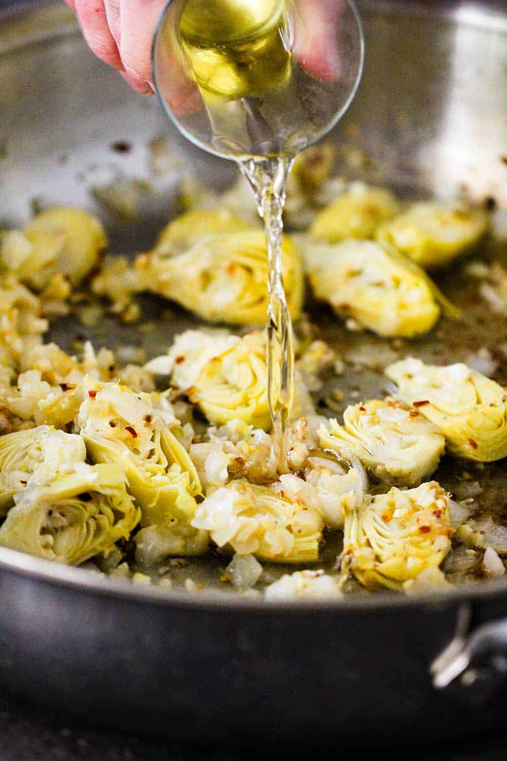 A hand pouring white wine from a small bottle into a skillet of cooked vegetables. 