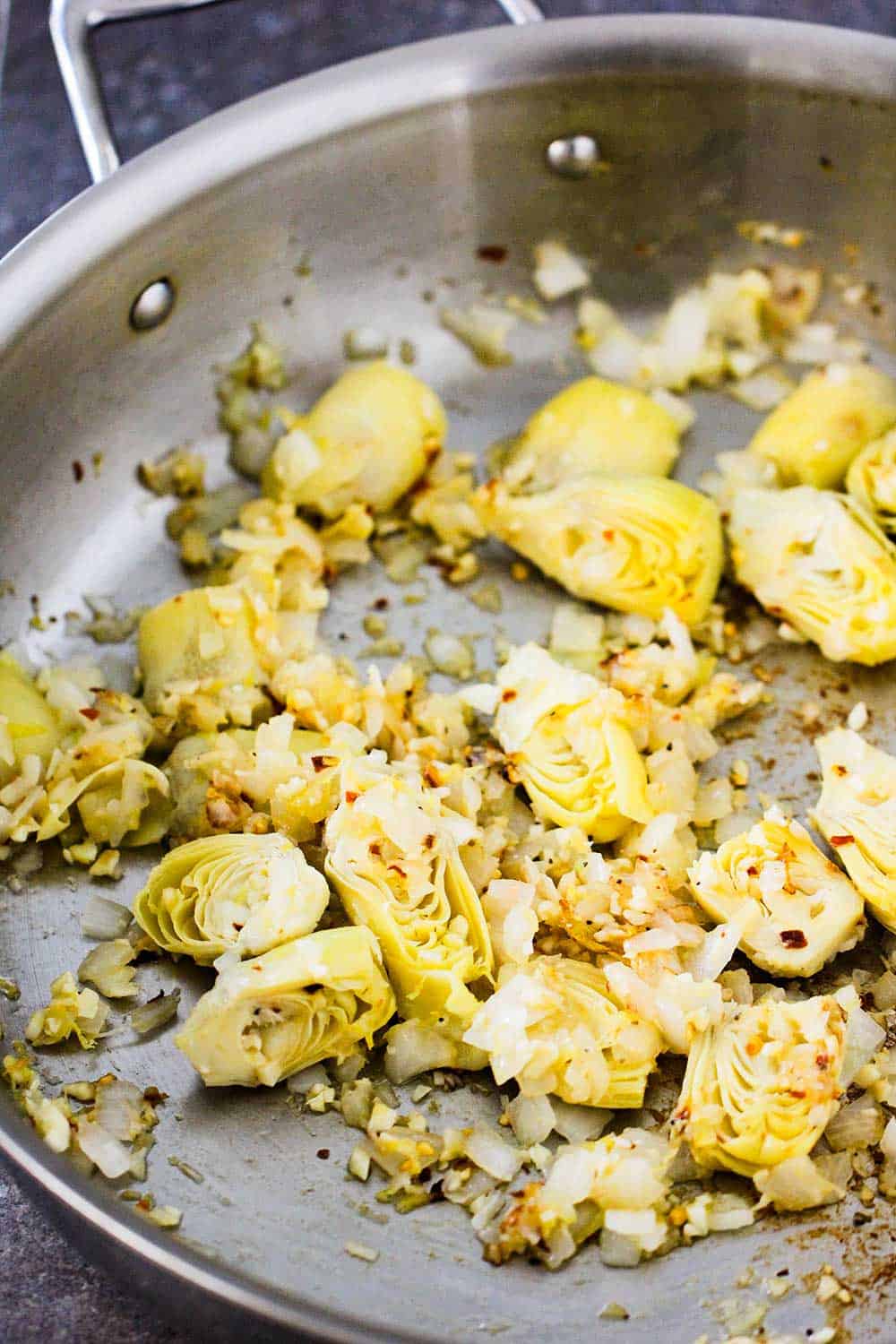 A large silver saucepan filled with sautéed artichokes, onions, and garlic. 