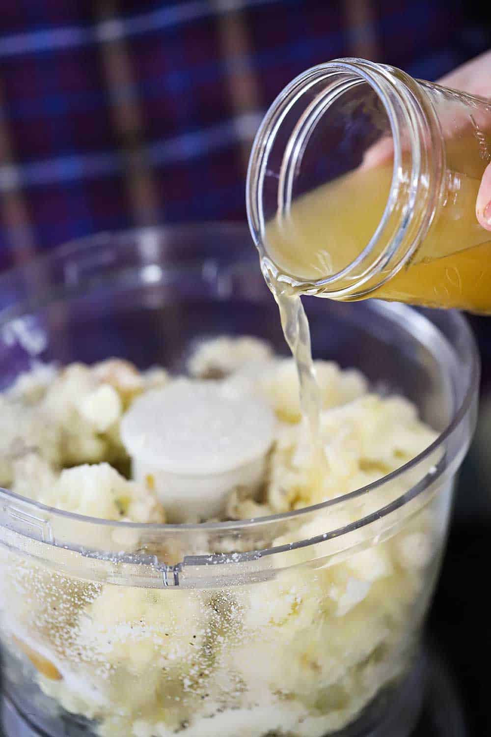 A hand pouring vegetable broth from a Mason jar into a food processor filled with steamed cauliflower florets and roasted garlic.