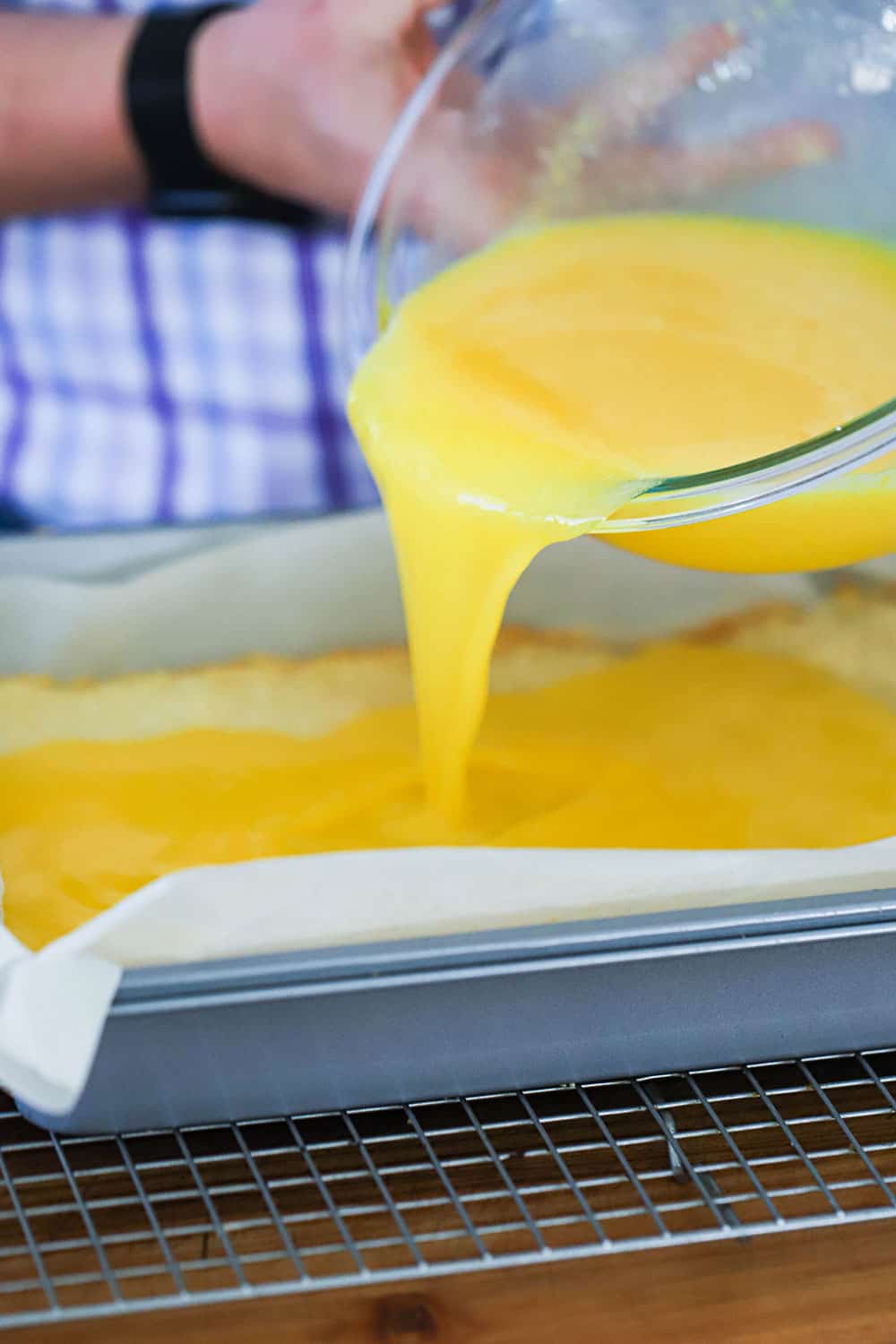 A person pouring lemon curd from a glass bowl into a metal baking pan lined with parchment paper. 