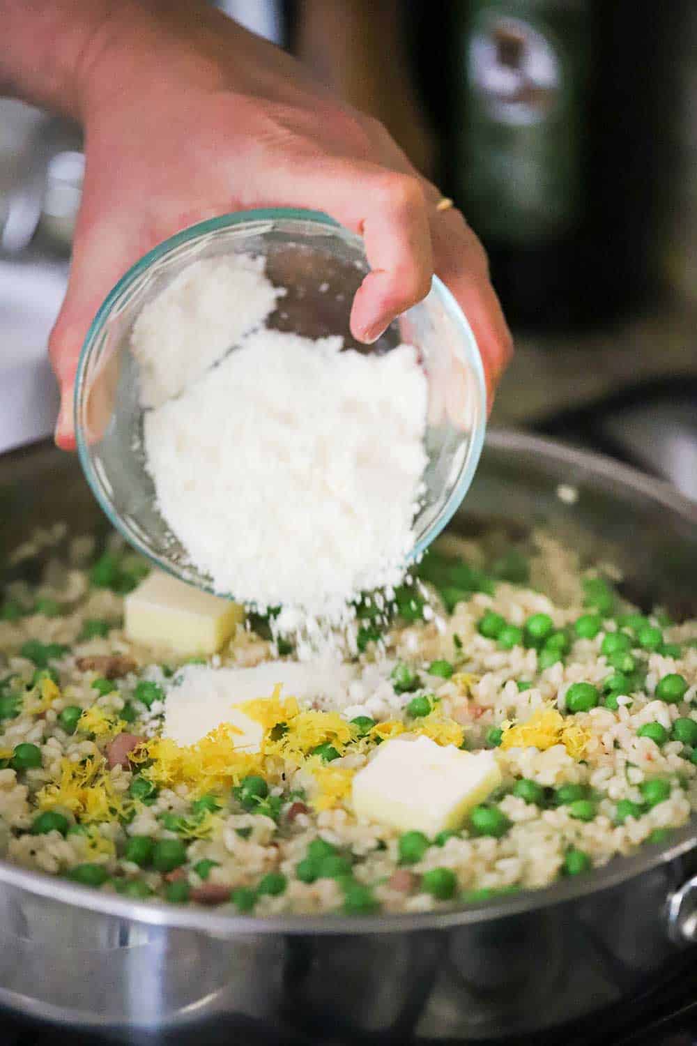 A person using their hand to transfer grated Parmesan cheese from a small bowl into a skillet of cooked risi e bisi. 