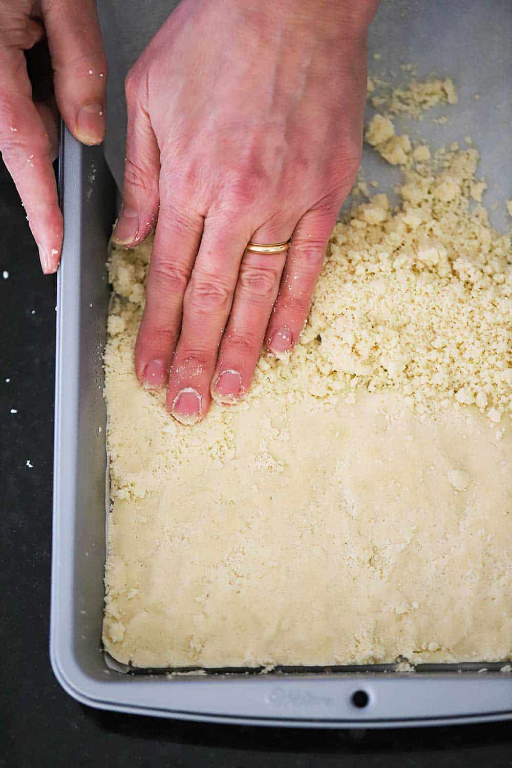 A person using his fingers and hand to press a shortbread dough into the bottom of a metal baking pan. 