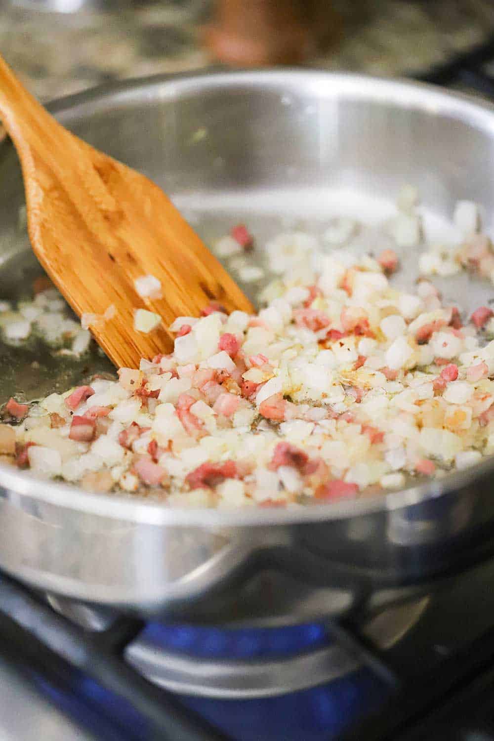 A large stainless steel skillet filled with sautéd chopped onions and diced pancetta and wooden spatula in the middle of it all. 