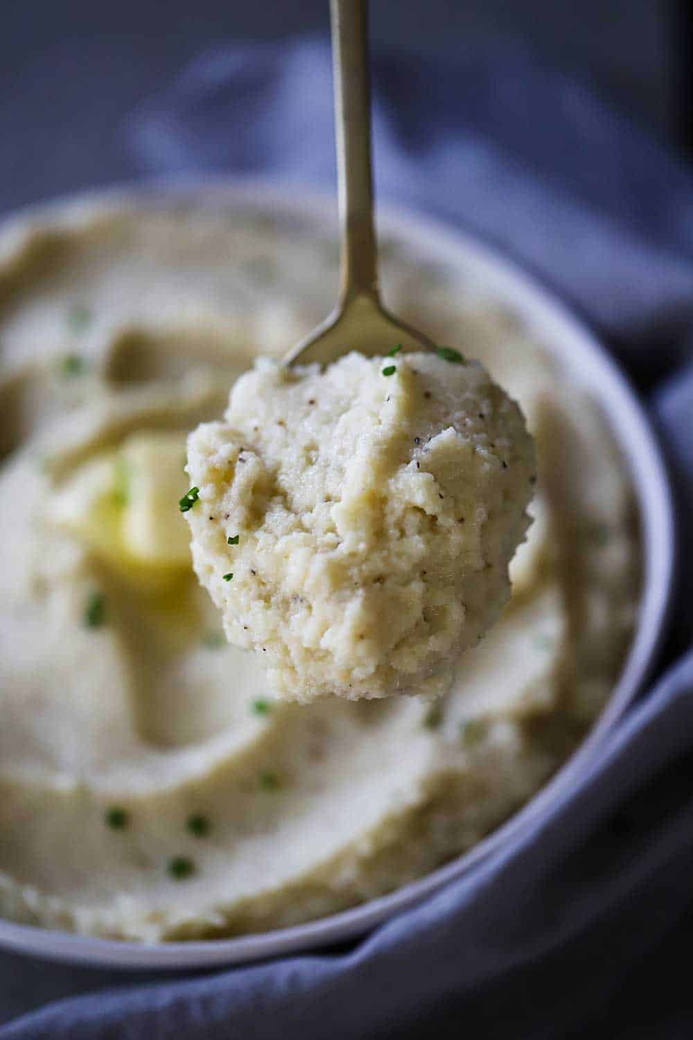 A serving spoon being lifted up filled with a helping of mashed cauliflower and roasted garlic over a bowl filled with the same. 