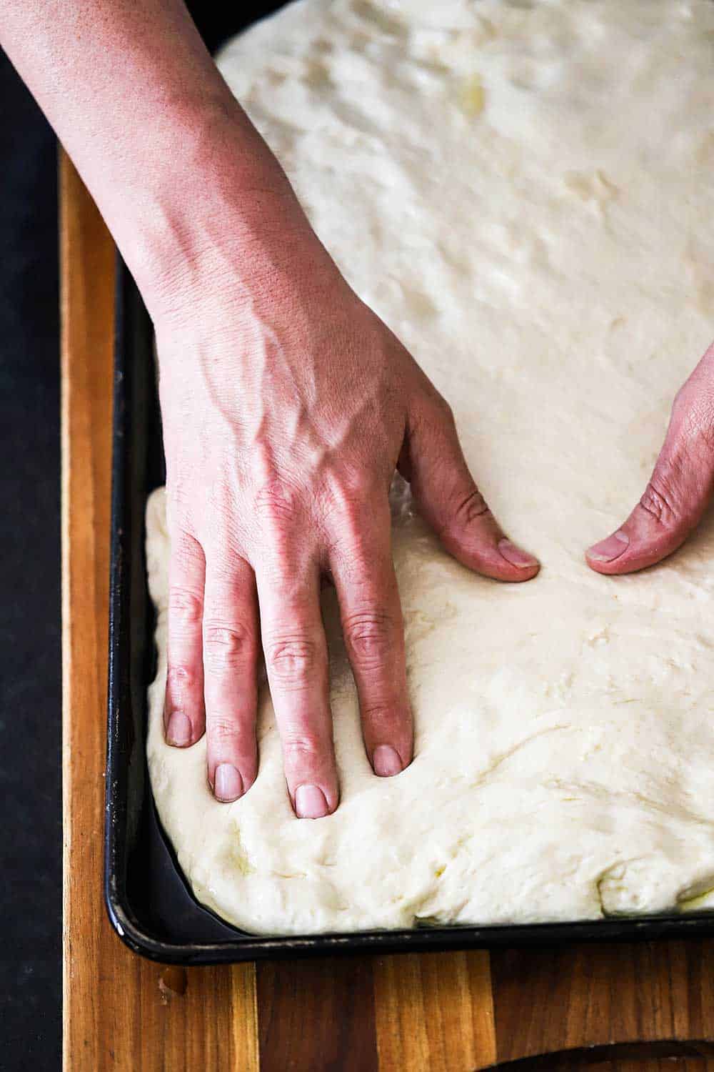 A pair of hands stretching focaccia dough into a rimmed baking pan. 