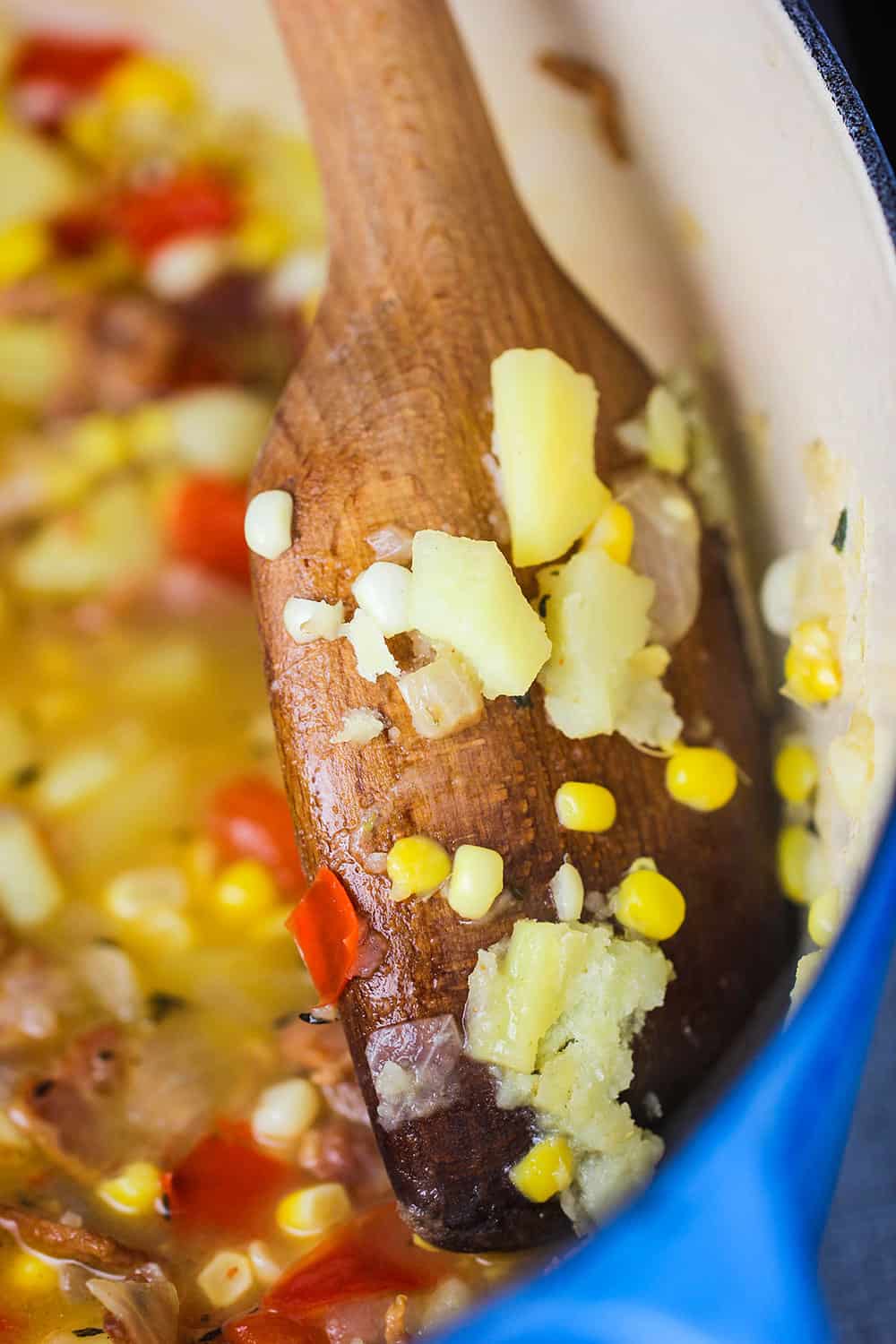 A wooden spoon smashing cooked potatoes and corn on the side of a blue Dutch oven for corn chowder. 