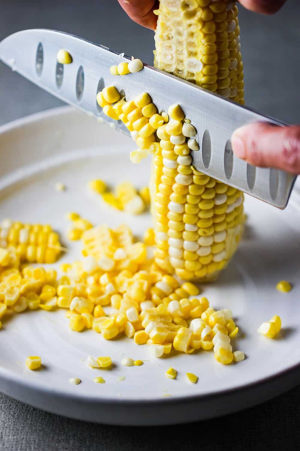 A hand using a large knife to remove corn kernels from an ear of corn. 