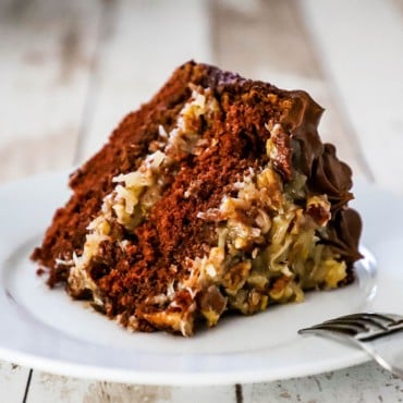 A large slice of German chocolate cake sitting on a white dessert plate with a fork next to it.